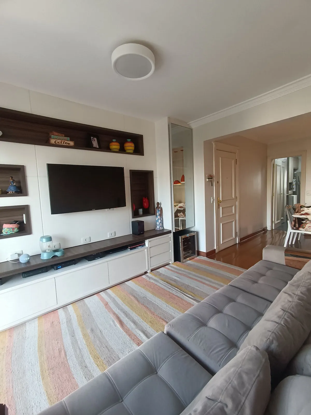 Sala de estar com painel branco, nichos amadeirados, TV, tapete listrado pastel e sofá cinza, antes da reforma