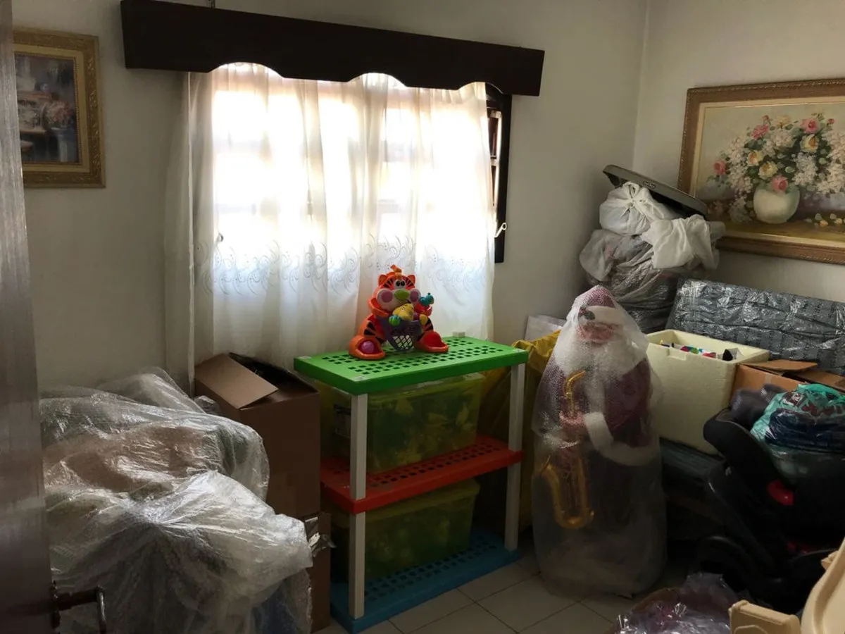 Quarto virado depósito, com bandô escuro na janela, caixas empilhadas e quadros de flores, antes da reforma