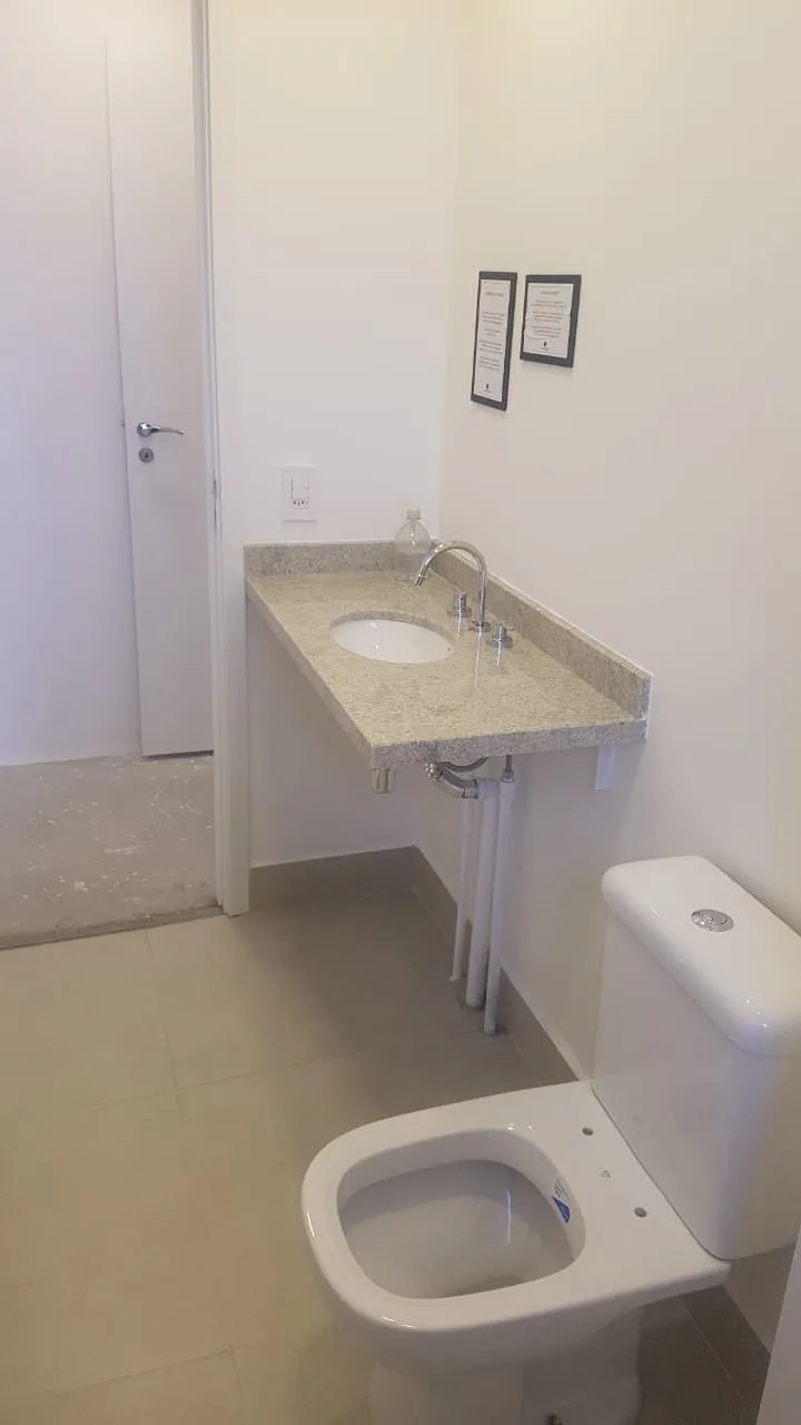 Banheiro com bancada de granito bege, cuba oval e vaso sanitário, piso e paredes claros, antes da reforma