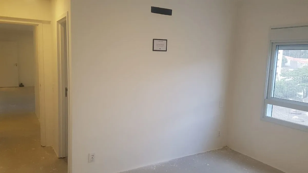 Quarto vazio de paredes brancas com porta para o corredor e janela lateral, piso de contrapiso, antes da reforma