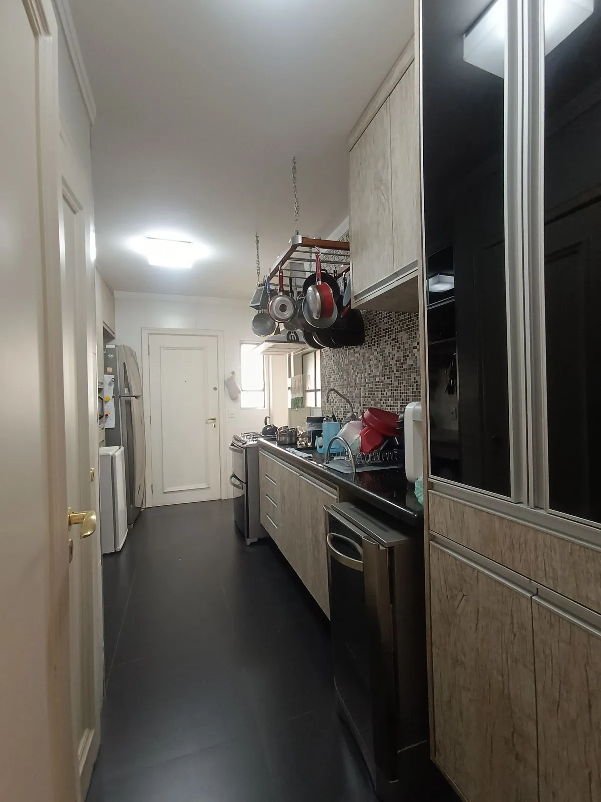 Cozinha estreita com marcenaria amadeirada, piso preto, varal de panelas, geladeira e fogão inox, antes da reforma