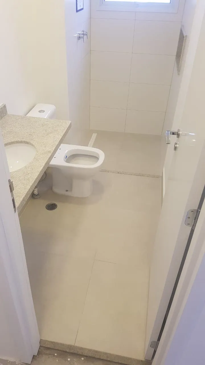Banheiro estreito com bancada de granito, vaso sanitário, box de vidro e piso bege, antes da reforma