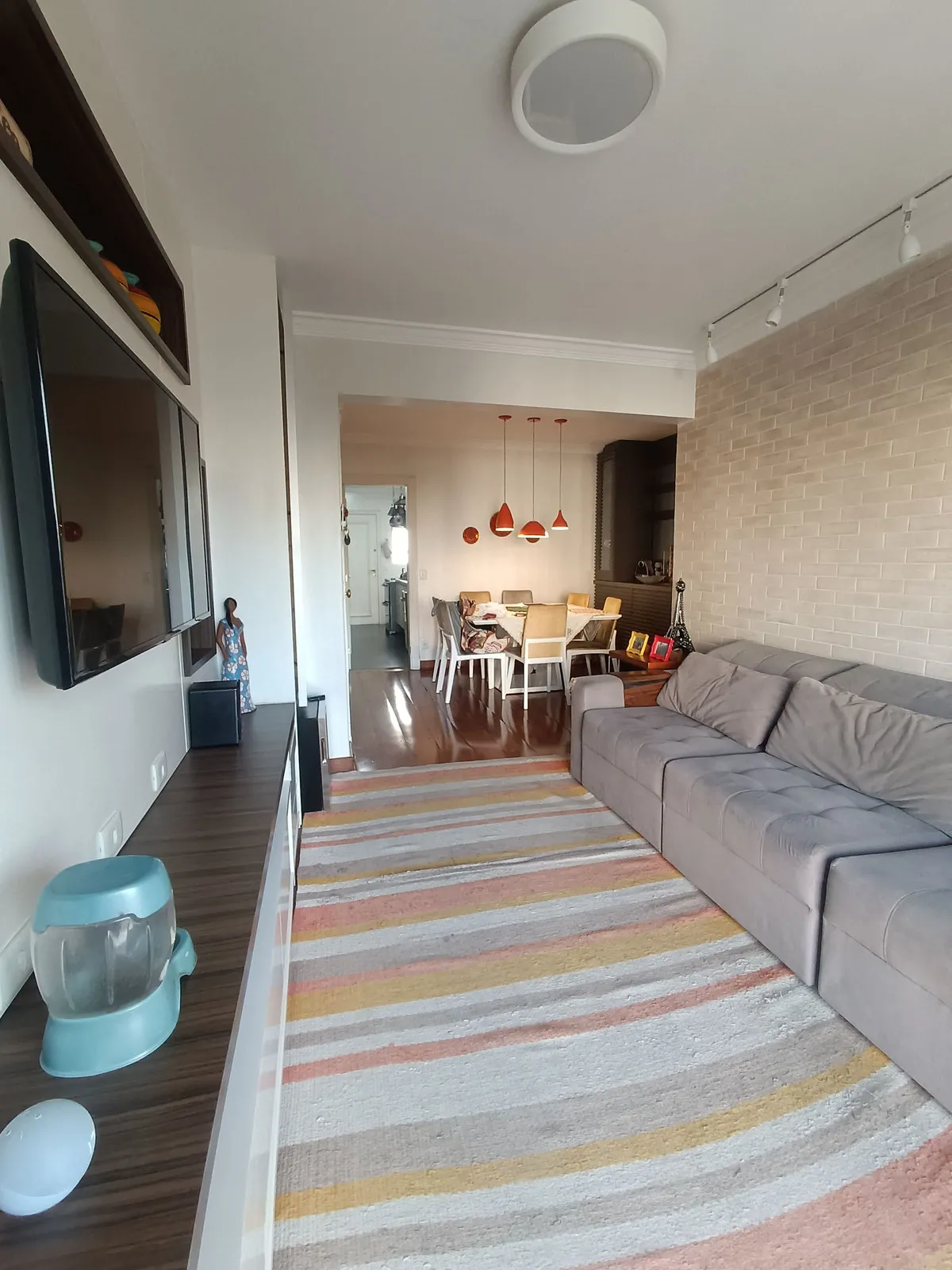 Sala alongada com sofá cinza, tapete listrado, parede de tijolinho claro e jantar ao fundo, antes da reforma