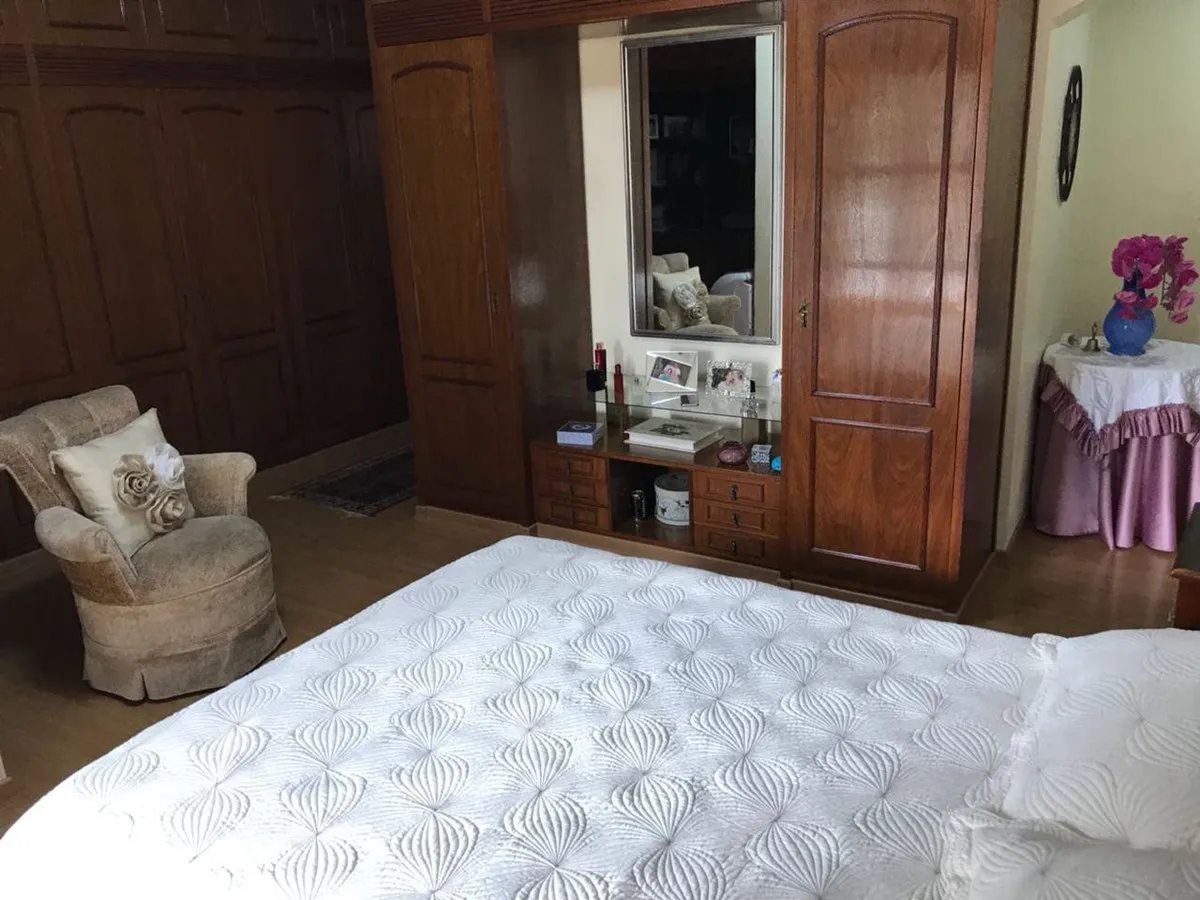 Suíte do casal com cama de colcha branca, parede de armários de madeira com penteadeira e espelho, antes da reforma