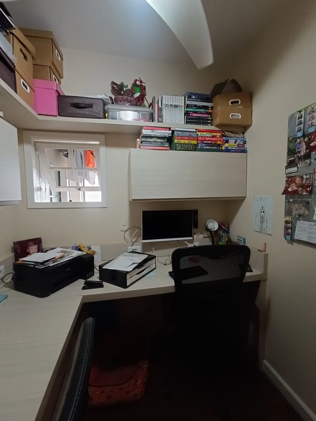Home office apertado com bancada branca em L, armários, prateleiras cheias e cadeira preta, antes da reforma