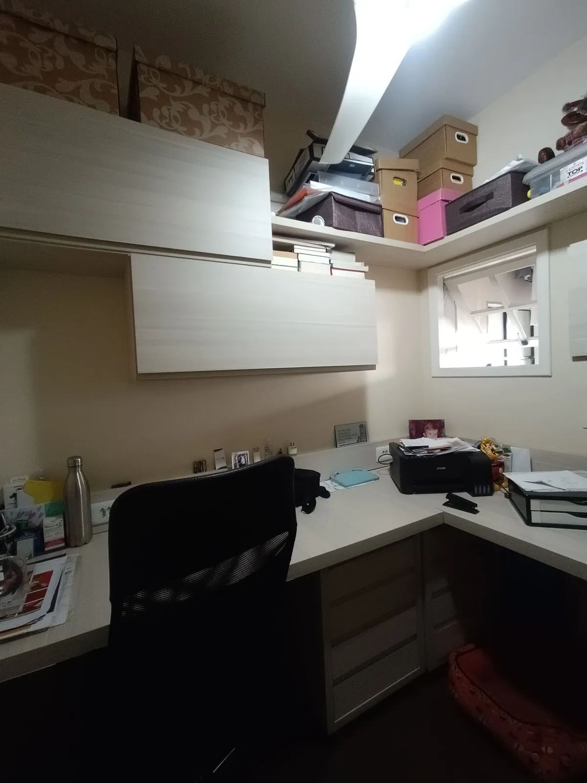 Home office em L com bancada clara, armários abarrotados de caixas, cadeira preta e janela interna, antes da reforma