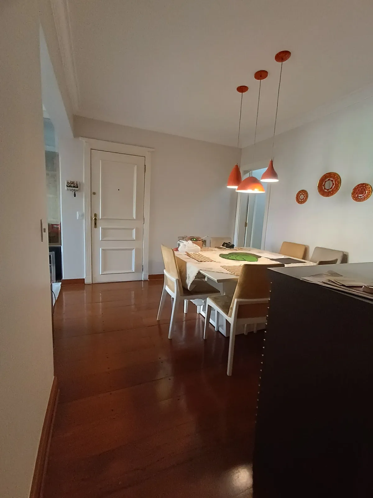 Sala de jantar com mesa branca, cadeiras claras, pendentes laranja e porta de entrada, antes da reforma