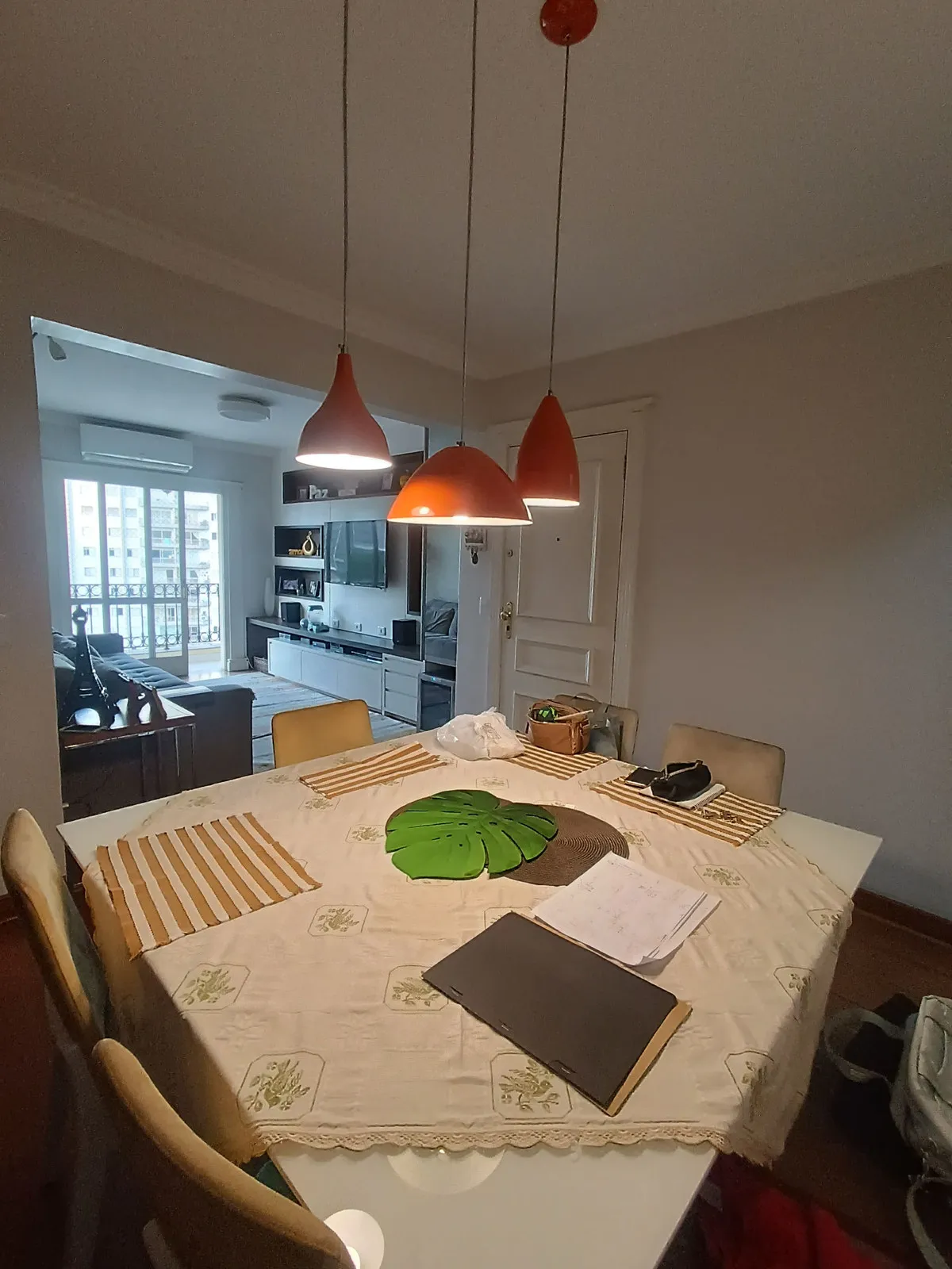 Mesa de jantar posta sob pendentes laranja, com sala e sacada ao fundo, antes da reforma
