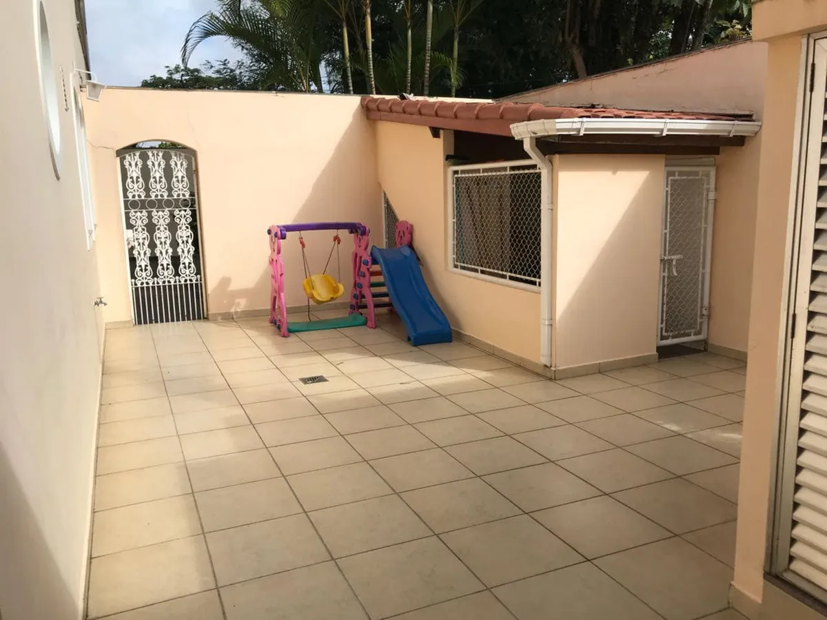 Quintal de piso cerâmico bege com balanço e escorregador infantil e portão de ferro, antes da reforma