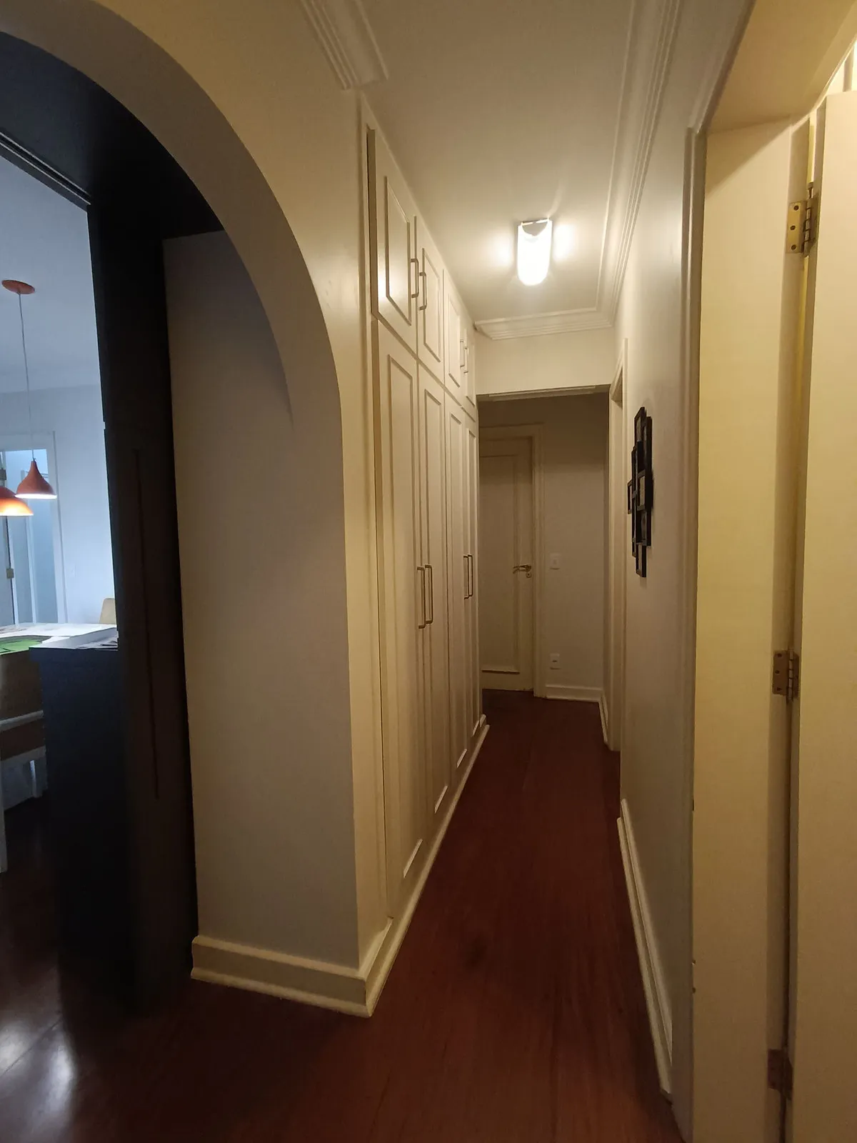 Corredor com armários brancos almofadados, arco de gesso e piso de madeira avermelhada, antes da reforma