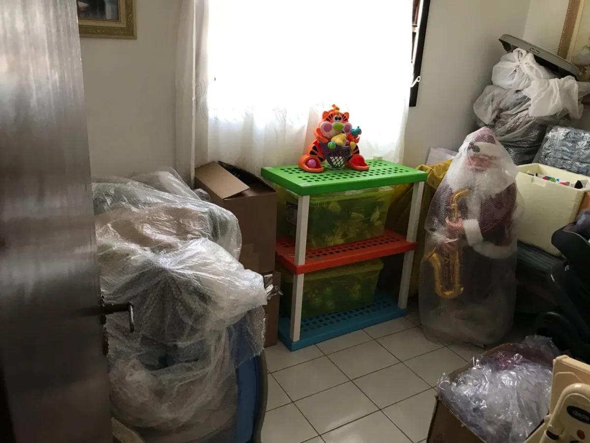 Cômodo usado como depósito, cheio de caixas, brinquedos e objetos embalados, com piso branco, antes da reforma