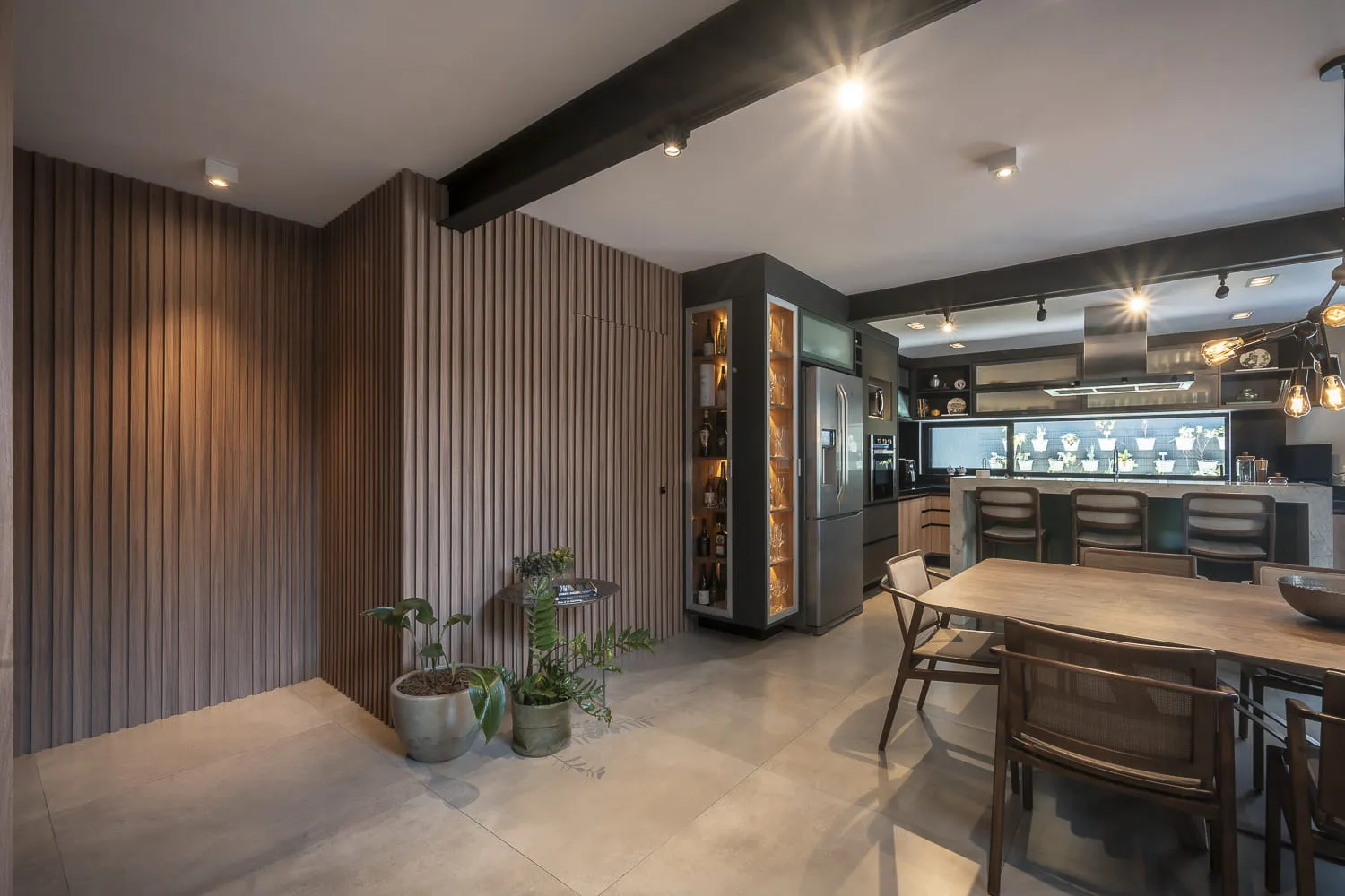 Sala de jantar com parede ripada de madeira, adega iluminada, geladeira inox e mesa de madeira ao lado da cozinha
