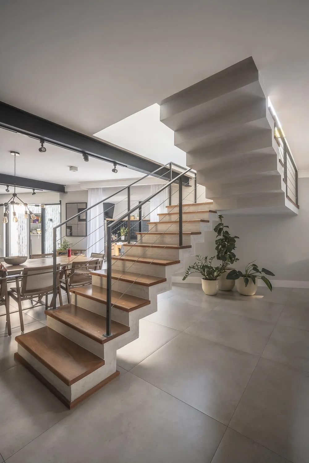 Escada de concreto com degraus de madeira e guarda-corpo de aço, plantas na base e sala de jantar ao lado