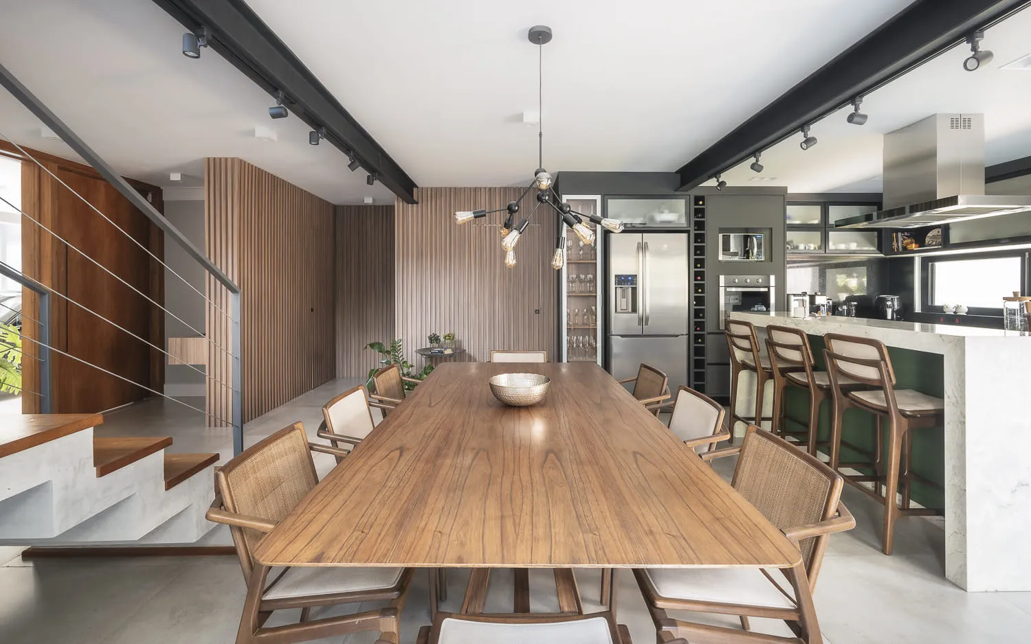 Mesa de jantar de madeira voltada para a cozinha, com península de mármore, banquetas verdes e escada ao fundo