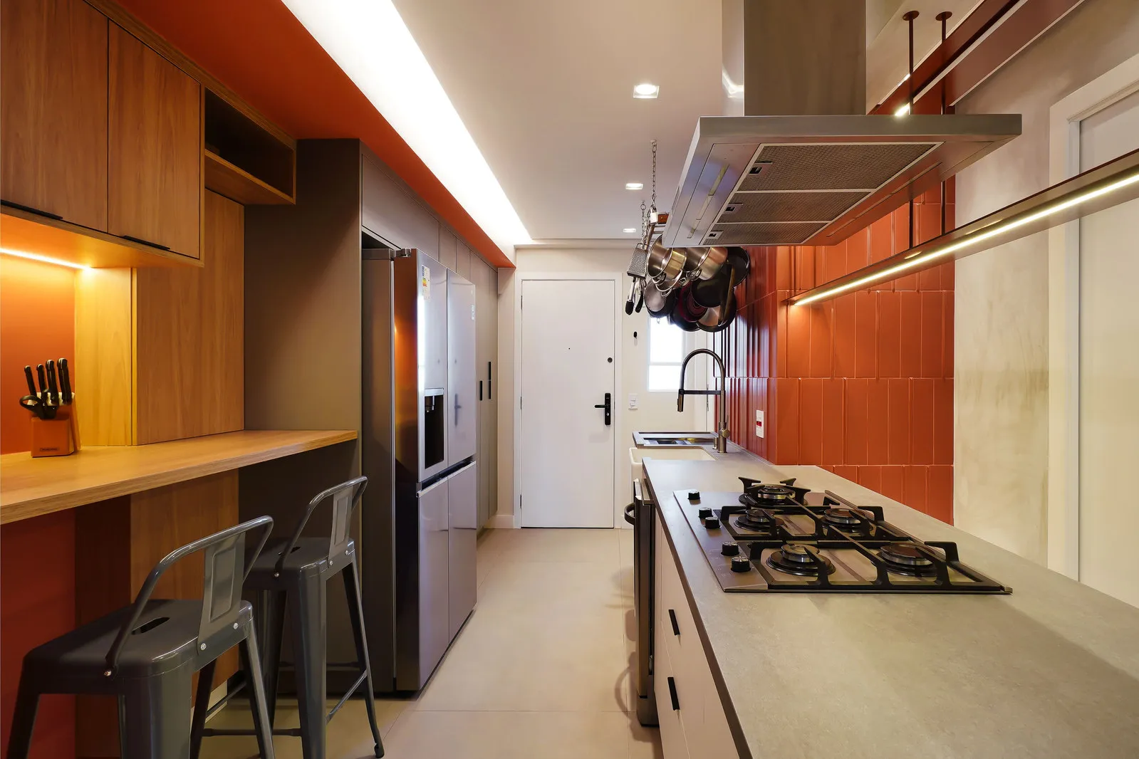 Cozinha corredor com parede de azulejo laranja, bancada de concreto, cooktop e varal de panelas suspenso