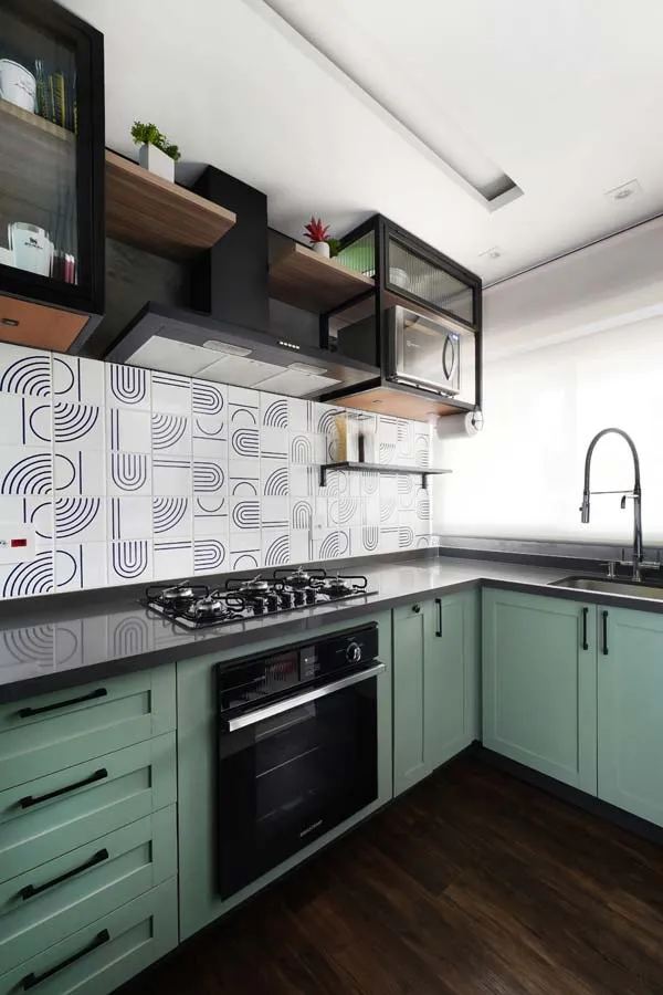 Cozinha em L com marcenaria verde-clara estilo Shaker, bancada cinza, cooktop e forno preto, revestimento geométrico e coifa preta