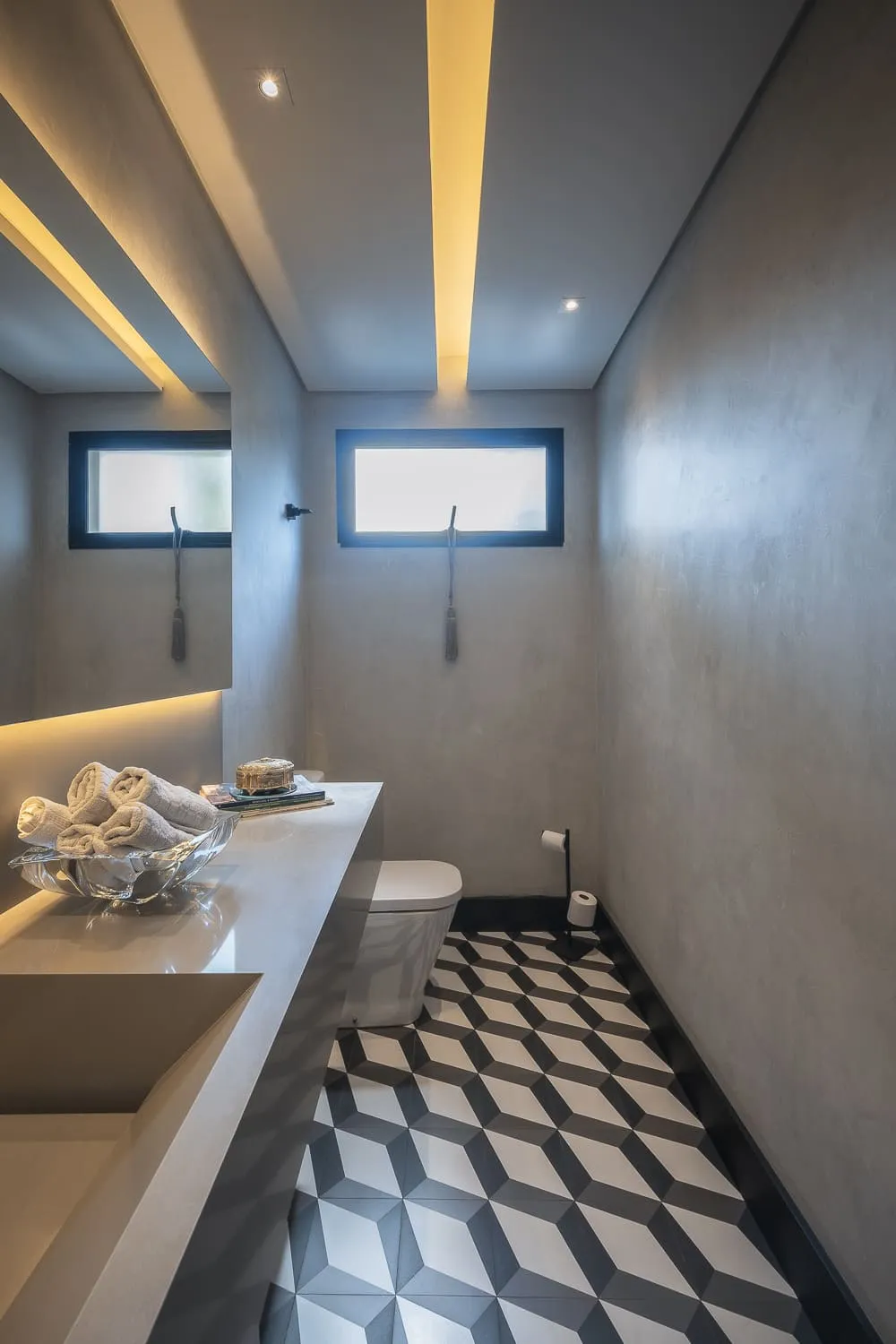 Lavabo de cimento queimado com bancada branca, janelas altas e piso de ladrilho geométrico em preto e branco