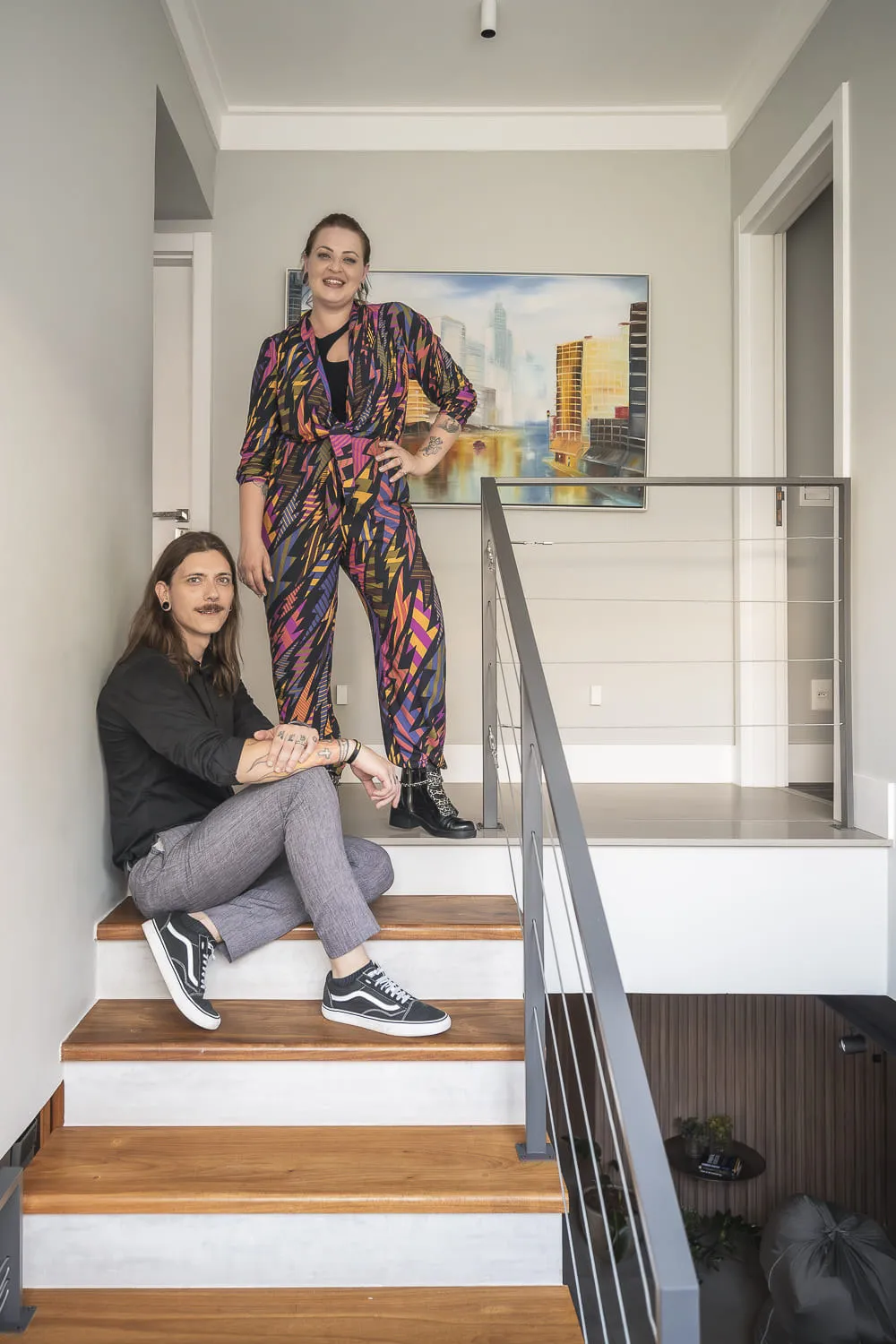 Deborah e Juliano, da Queen B, na escada de madeira do projeto, com quadro de paisagem urbana na parede ao fundo