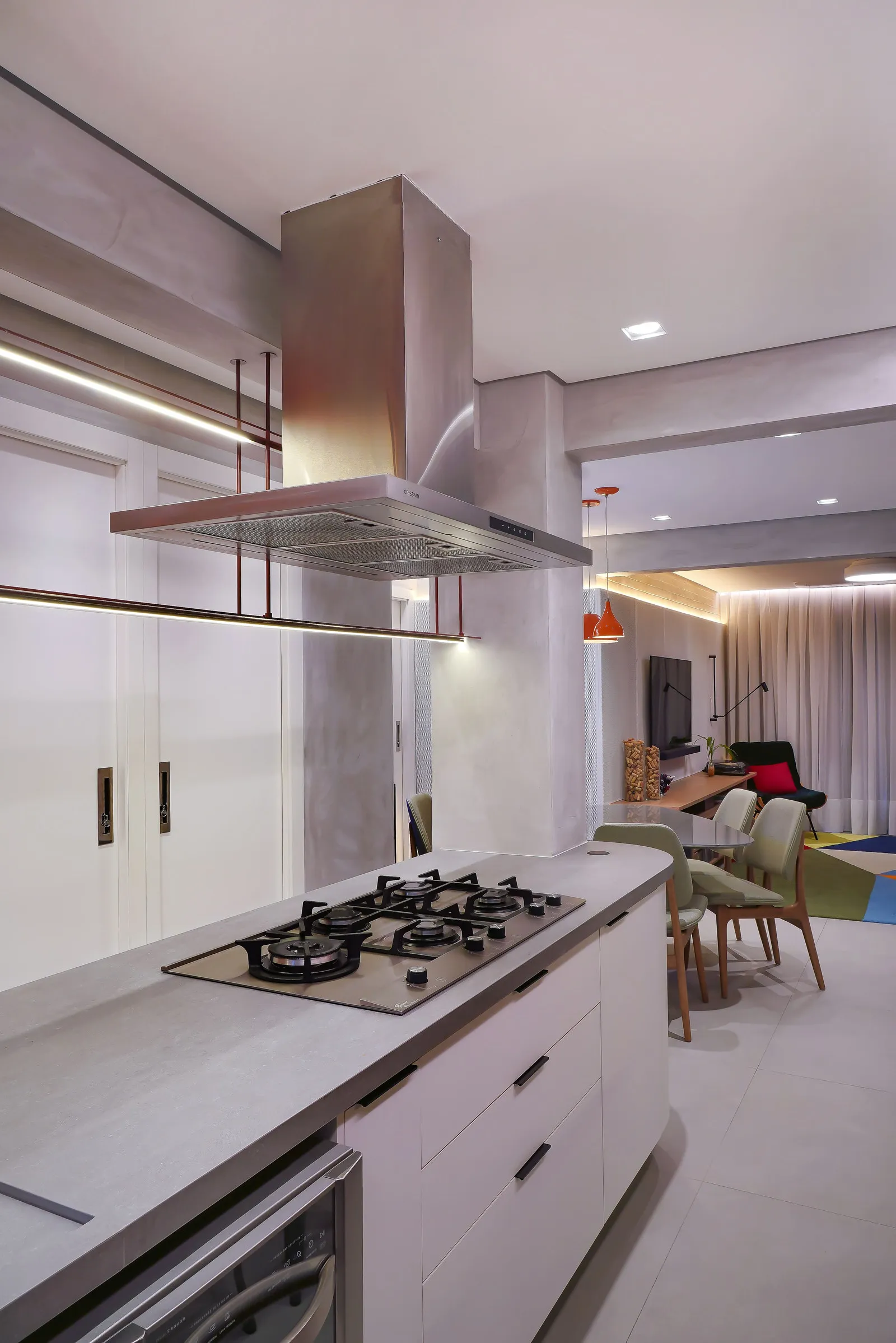 Ilha de cozinha com bancada de concreto, cooktop e coifa inox suspensa, aberta para a sala de jantar ao fundo