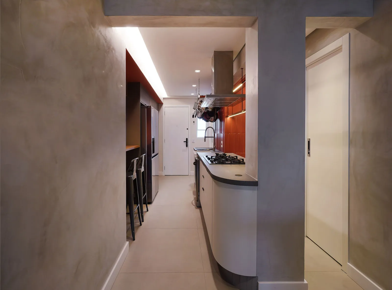 Corredor da cozinha com bancada de concreto arredondada, cooktop, azulejo laranja e porta de entrada ao fundo