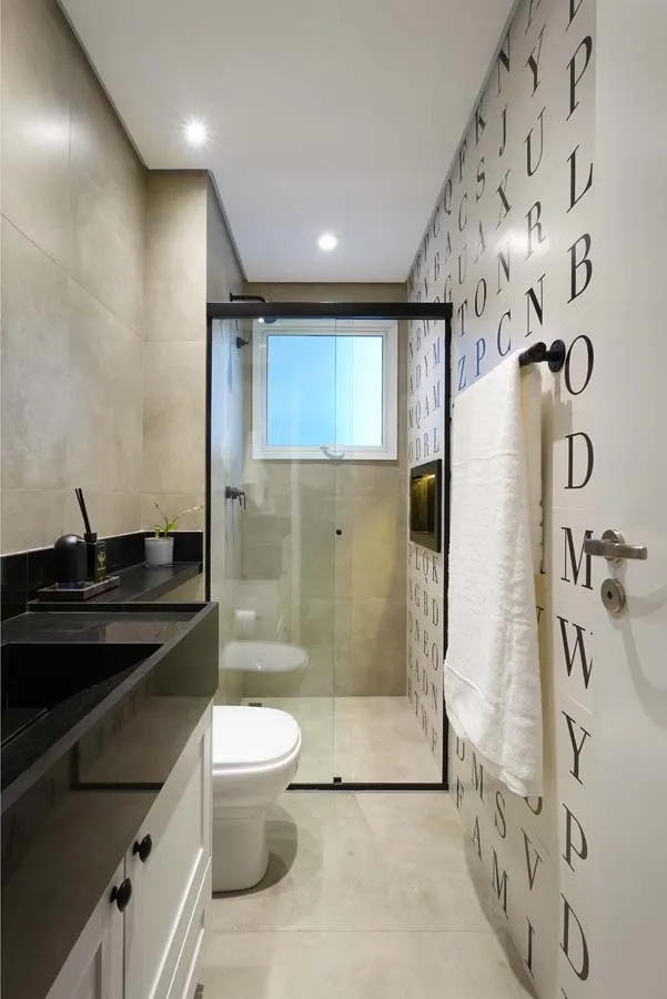Banheiro estreito com bancada preta e cuba esculpida, box de vidro com janela, parede de revestimento de letras e porcelanato bege