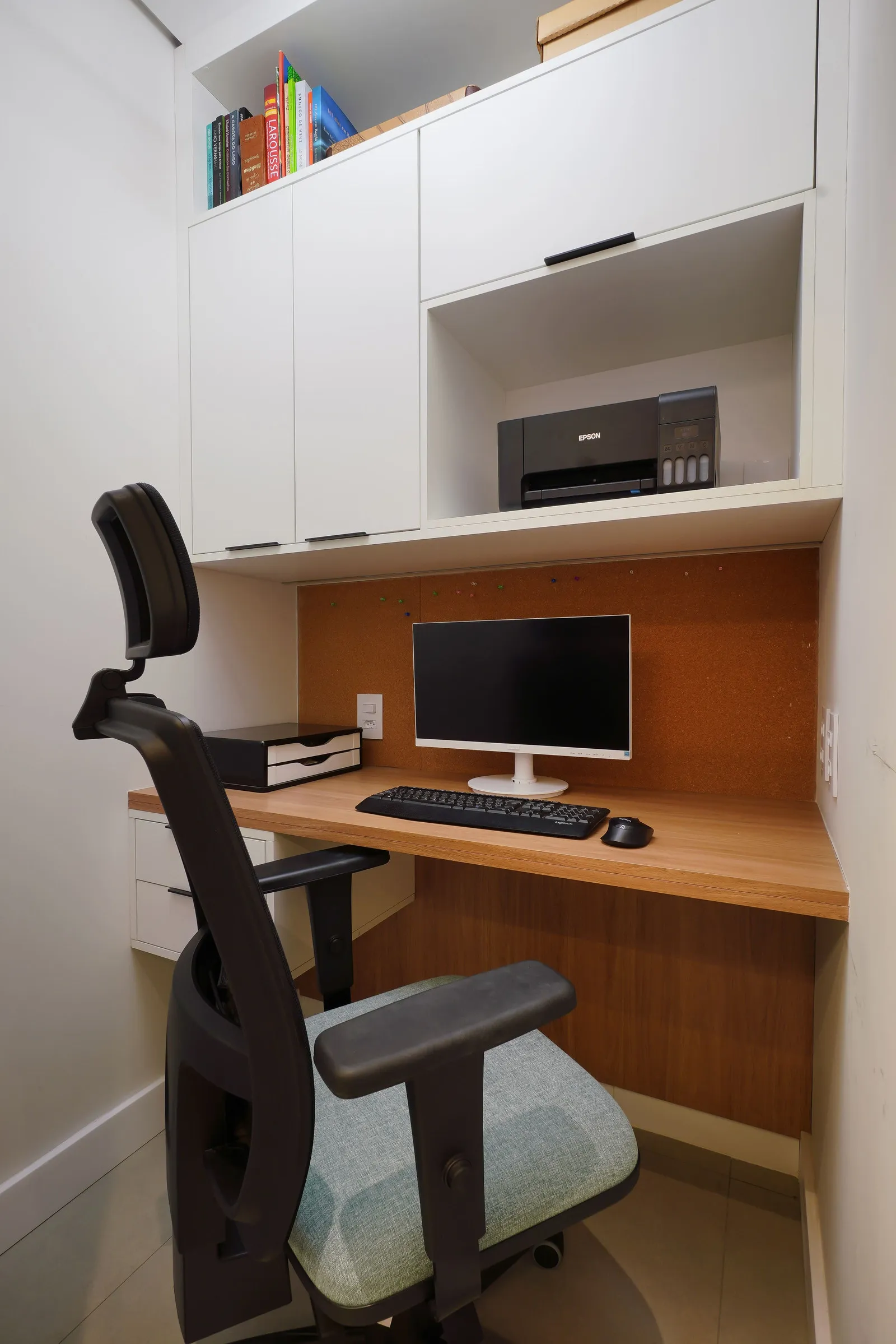 Home office compacto com bancada de madeira, armários brancos, quadro de cortiça, monitor e cadeira ergonômica