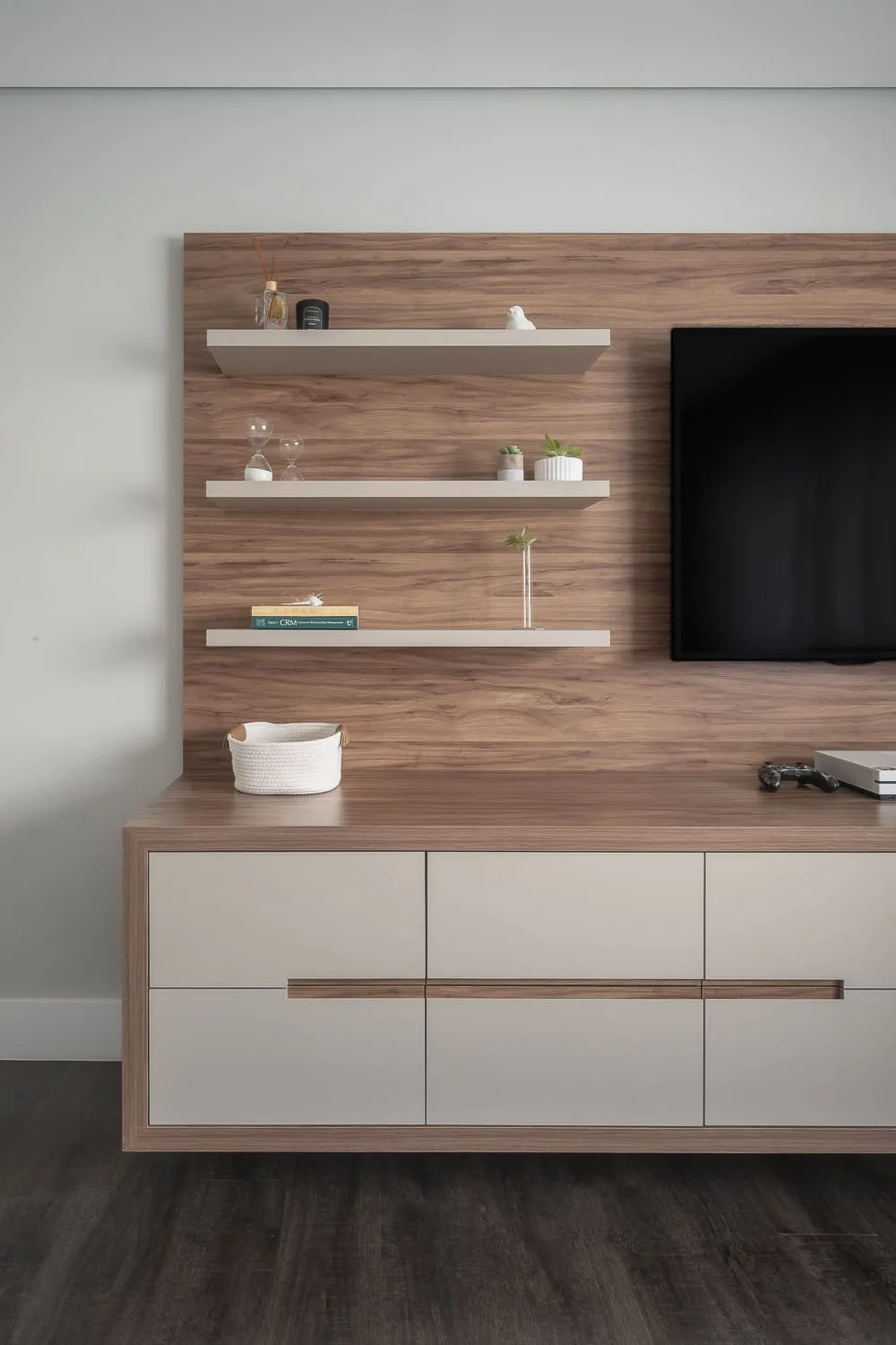Painel de TV amadeirado com prateleiras suspensas e rack de gavetas claras sobre piso de madeira escura