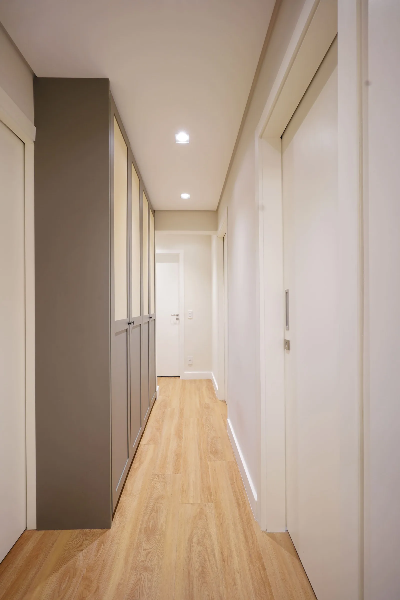 Corredor com piso laminado de madeira clara, armários embutidos em bege e fendi e portas brancas