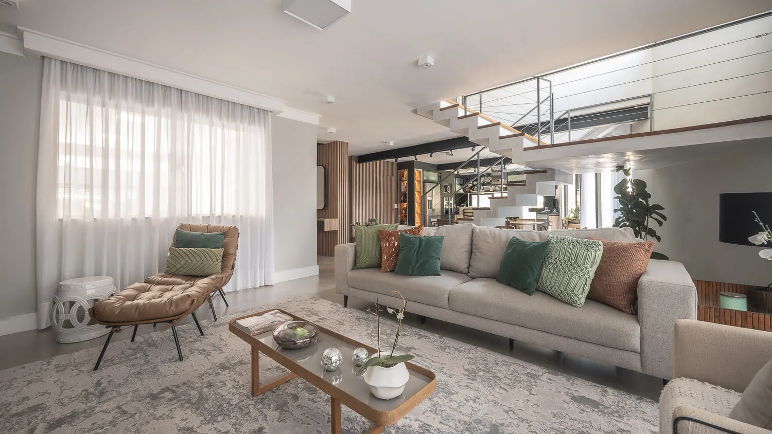 Living integrado com sofá cinza, chaise de couro caramelo e mesa de centro de madeira, sob escada de concreto e mezanino
