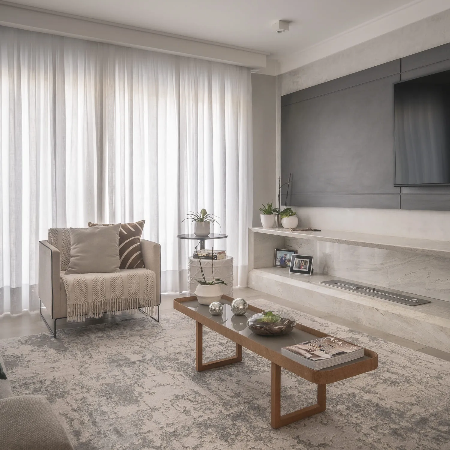 Sala de TV com poltrona bege, mesa de centro de madeira e painel cinza sobre bancada de mármore com lareira ecológica