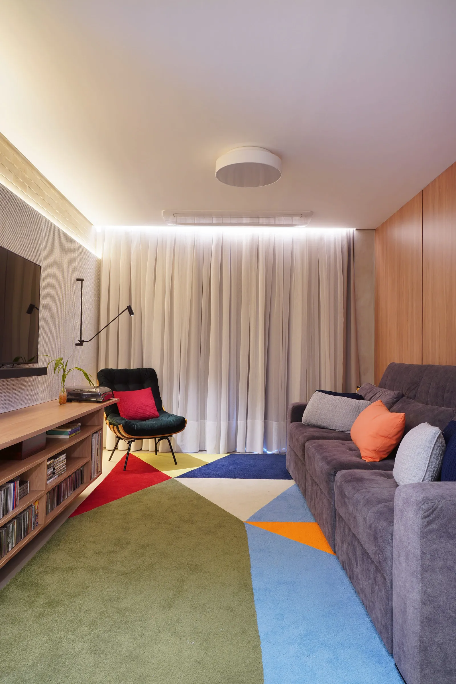 Sala de TV com sofá cinza, tapete geométrico colorido, poltrona Costela preta e rack de madeira com discos