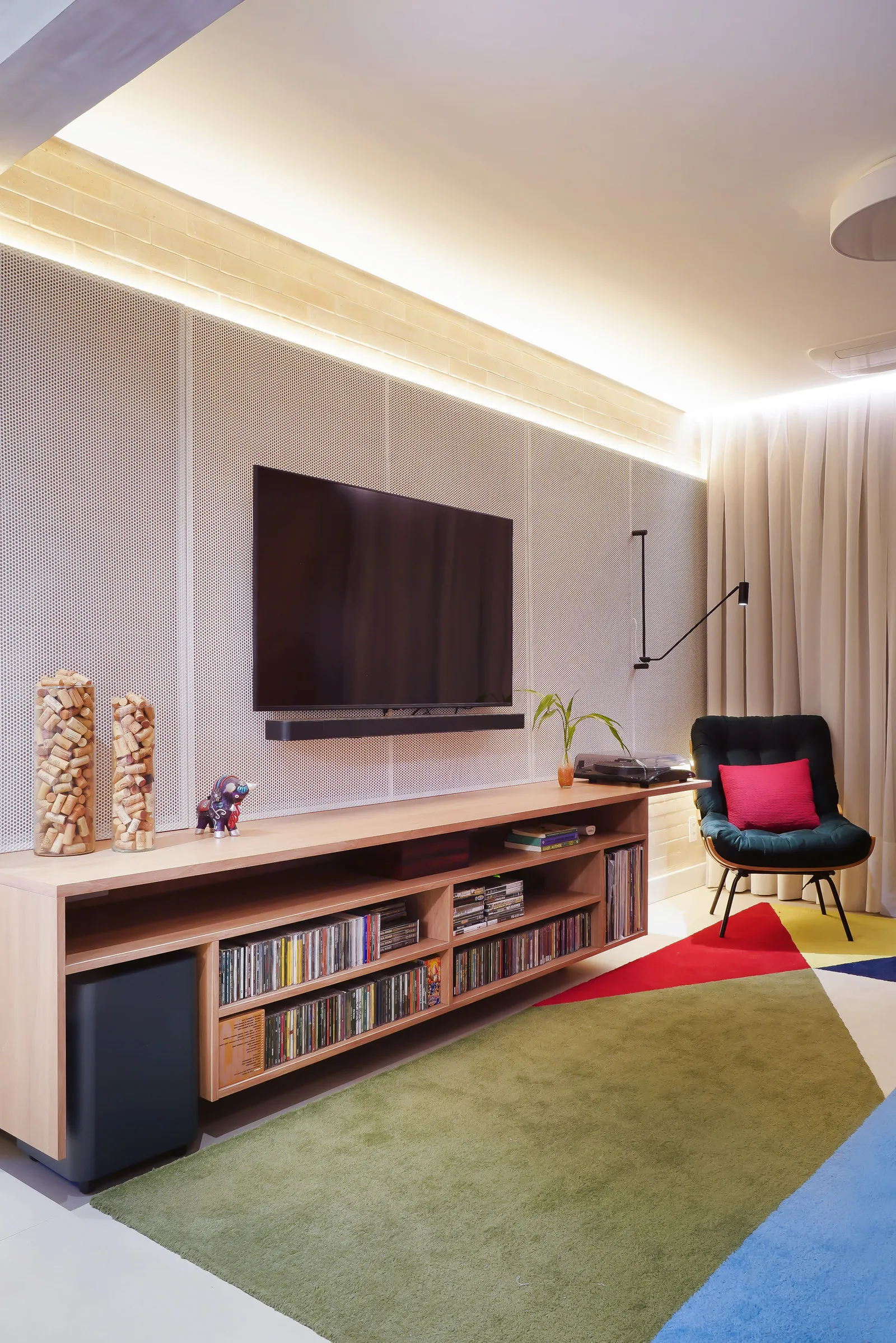 Painel de TV em metal perfurado branco com luz indireta, rack de madeira cheio de CDs e poltrona preta ao lado