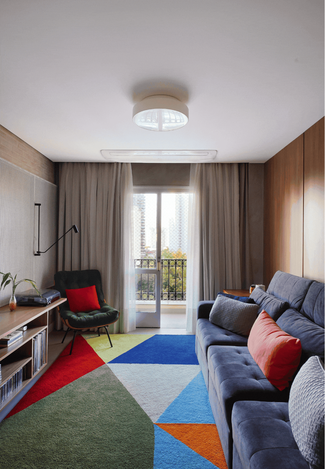 Sala de estar com sofá cinza, tapete geométrico colorido, poltrona preta e painel de madeira, com sacada ao fundo