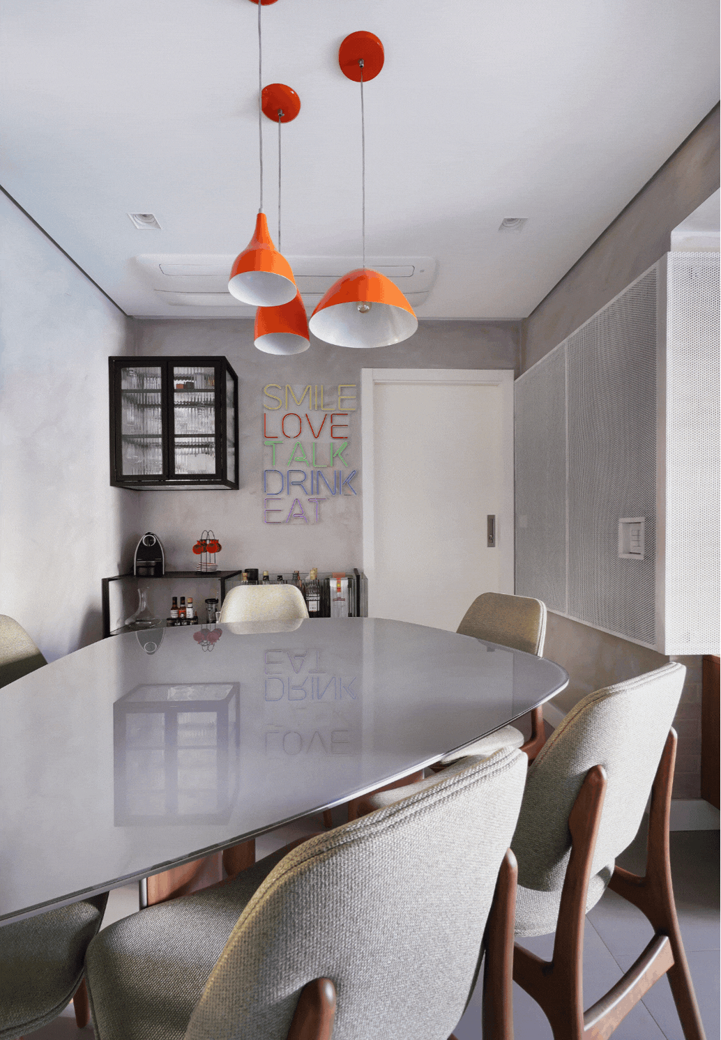 Sala de jantar com mesa oval de vidro, cadeiras estofadas, pendentes laranja e letreiro de neon na parede