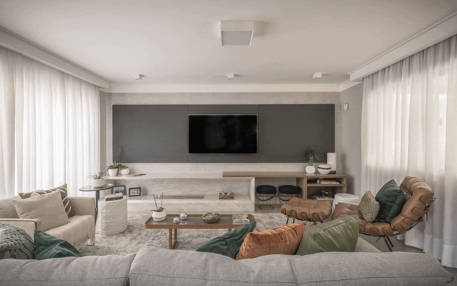 Sala de estar ampla com TV em painel cinza sobre bancada de mármore, sofás cinza e poltronas de couro caramelo