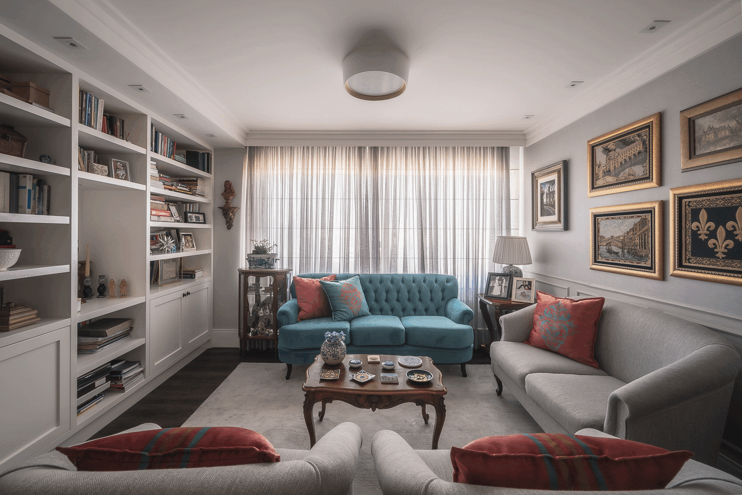 Sala de estar com sofá azul capitonê, sofás cinza, estante branca de livros e quadros dourados na parede