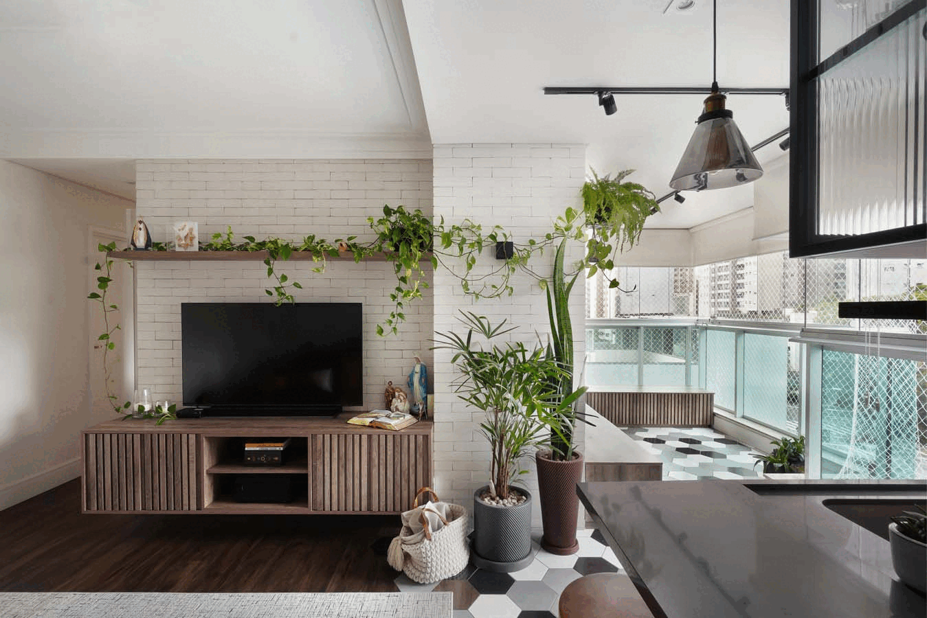 Sala de estar integrada à varanda, com TV no painel de tijolo branco, plantas pendentes, vasos de palmeira e piso hexagonal encontrando a madeira