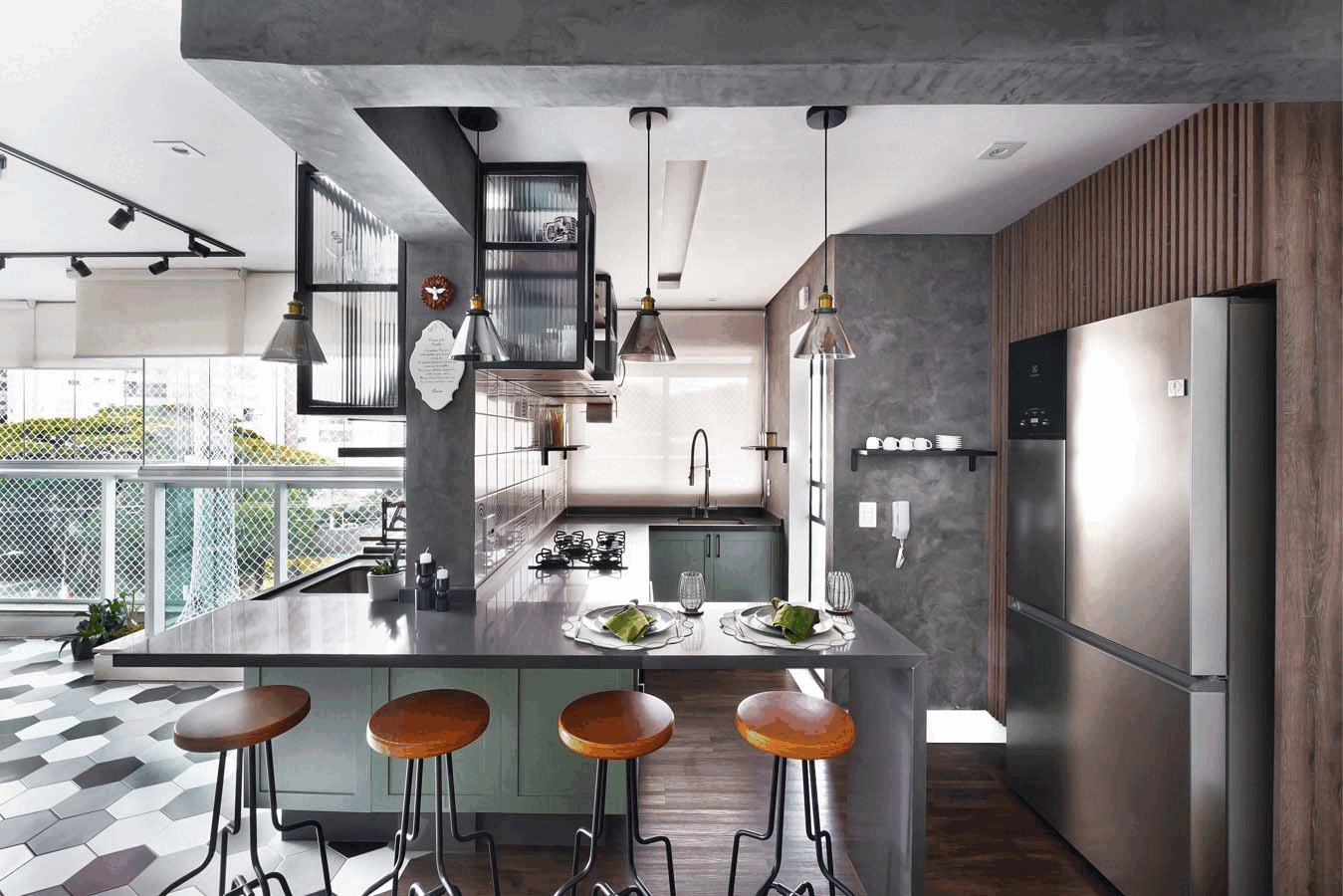 Cozinha ampla com teto de concreto e pendentes de vidro, península cinza posta, marcenaria verde e geladeira inox, com a varanda de piso hexagonal à esquerda