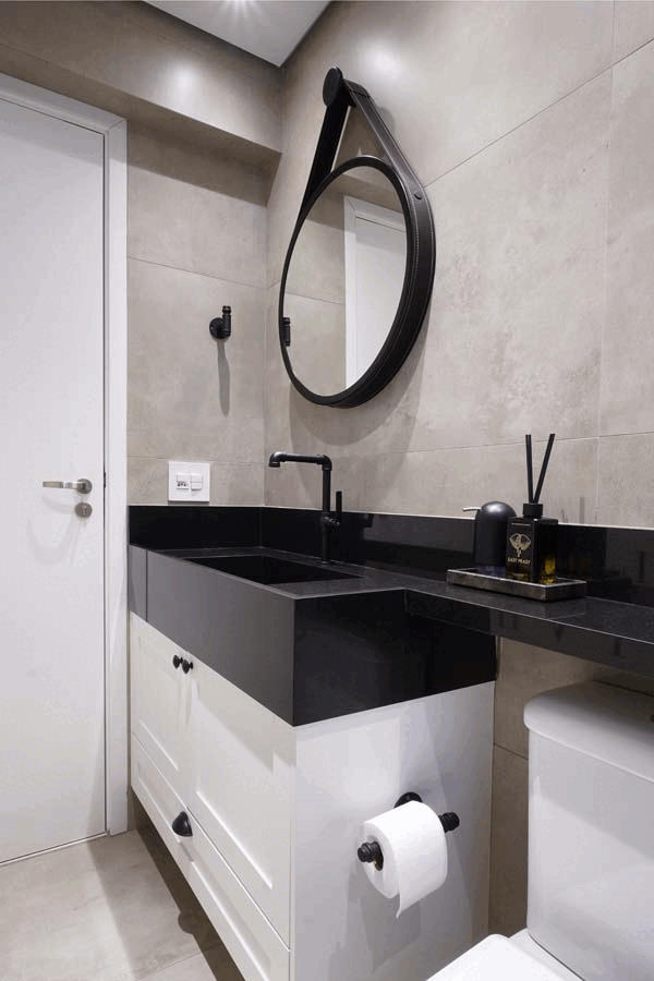 Lavabo com espelho redondo de alça preta, bancada de pedra preta com cuba esculpida e torneira preta, marcenaria branca e porcelanato bege