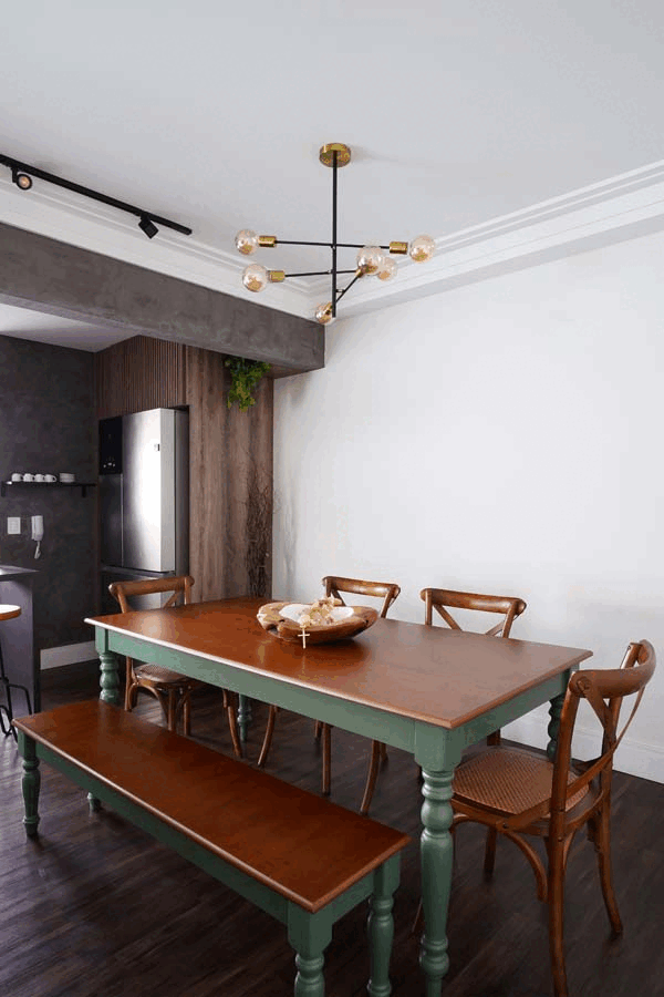 Sala de jantar com mesa de madeira de pés verdes, banco e cadeiras de madeira, lustre sputnik e painel ripado de madeira ao fundo com geladeira inox