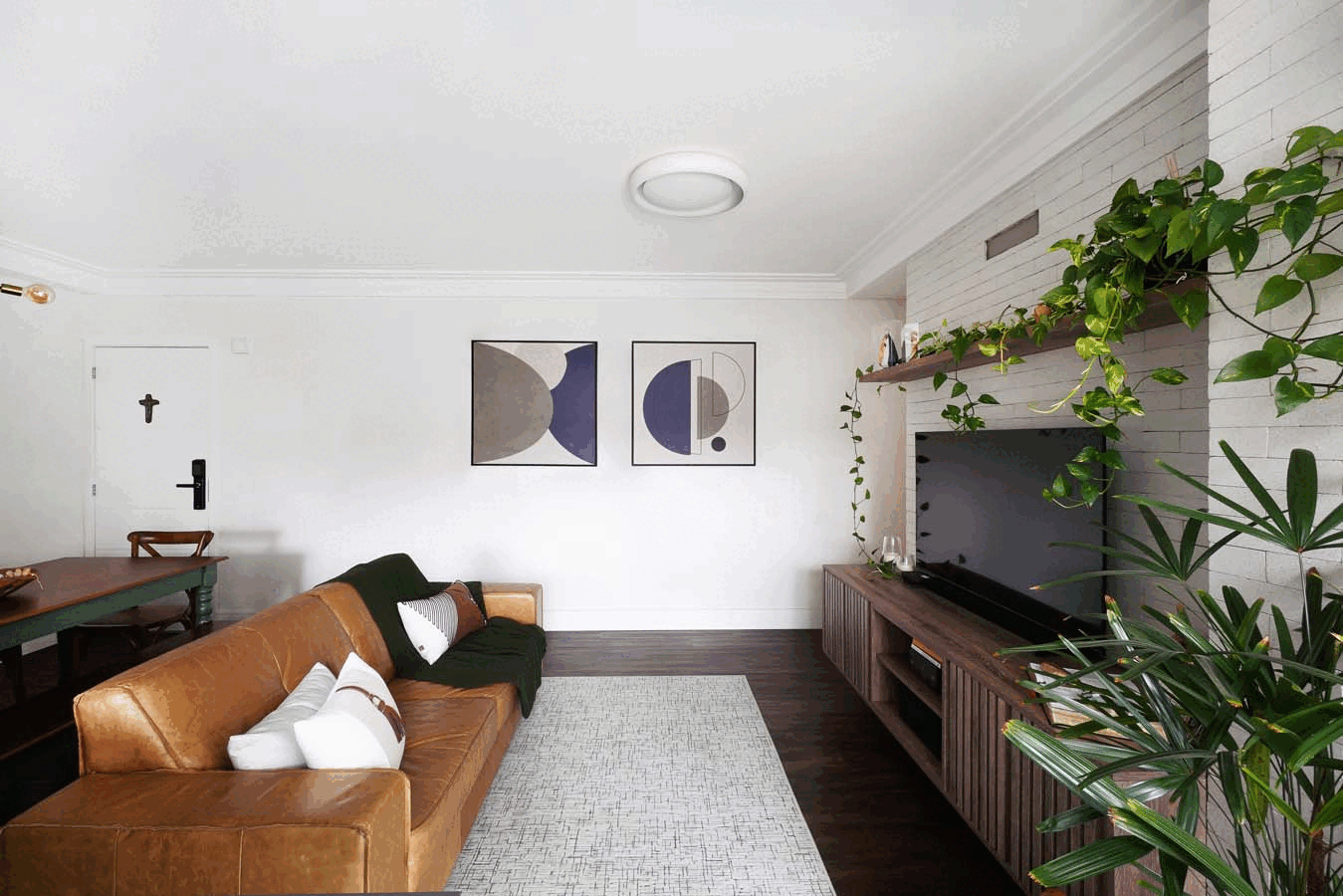 Sala de estar com sofá de couro caramelo, dois quadros geométricos na parede branca, TV em rack ripado com plantas pendentes e tapete claro sobre piso escuro