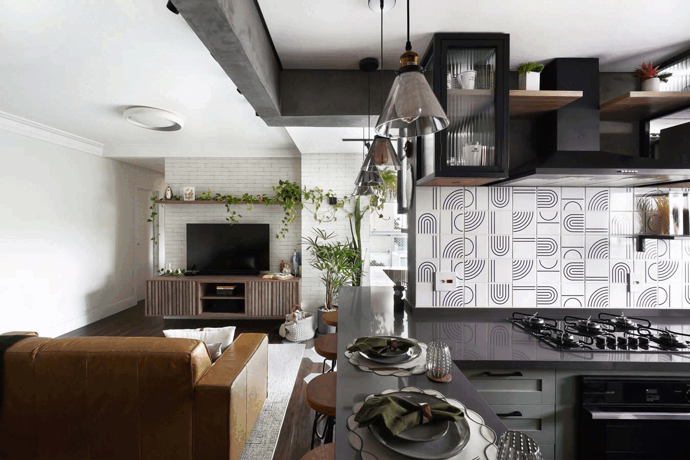 Cozinha com revestimento geométrico preto e branco, cooktop na ilha cinza, armários de vidro com moldura preta e pendentes, com a sala e o painel de tijolo ao fundo